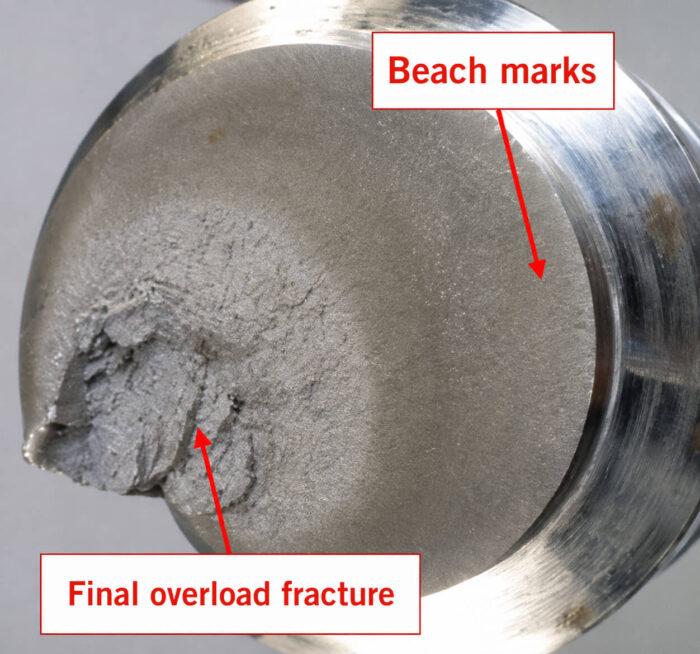 Fracture surface of a component showing beach marks and an adjacent final overload fracture area, both labeled with red arrows
